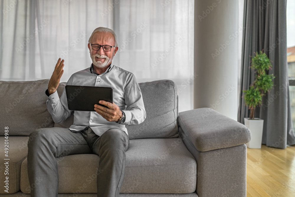 Smiling cheerful happy senior man waving and gesturing to camera having online video call on hand held tablet portable device talking to and connecting with family or friends