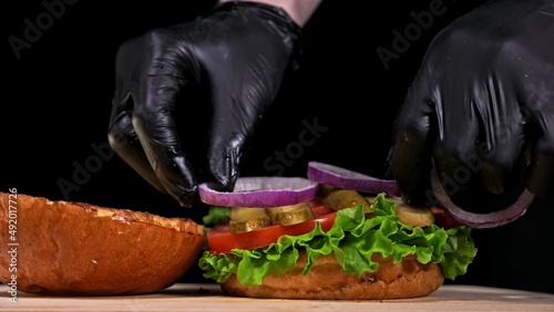 Craft burger is cooking on black background in black food gloves. Consist: sauce, lettuce, tomato, red onion, pickle, cheese, bacon, air bun and marbled meat beef. Not made ideal. Looks real, loving