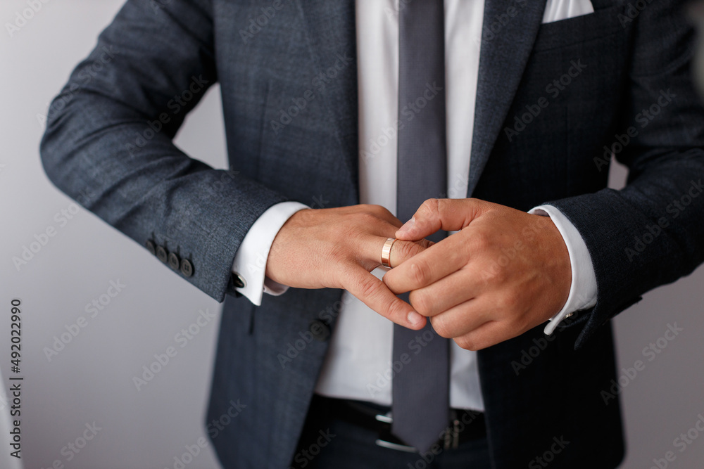 Obraz premium stylish adult male groom in a dark jacket with a tie and in a white shirt puts a golden ring on his finger on a gray background, without a face