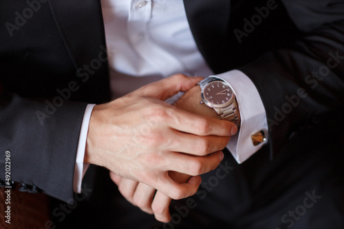 a man in a black classic tuxedo jacket and a white shirt holds a steel classic mechanical swiss watch