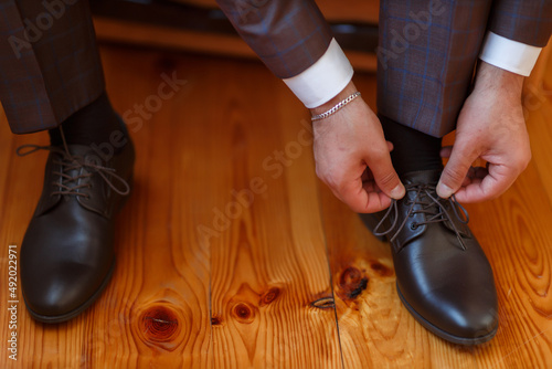 stylish man in classic brown suit tying shoelaces on brown leather shoes on wooden parquet