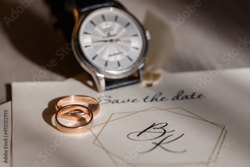 gold wedding rings lie on top of each other on an invitation card near a classic silver mechanical men's watch, close-up