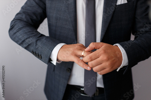 stylish adult male groom in a dark jacket with a tie and in a white shirt puts a golden ring on his finger on a gray background, without a face