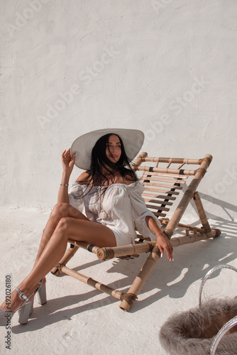 vacation mood. brunette woman with long legs in white summer dress and white hat sits on a beach chair and looks away with smile near white wall background. greek villa. travel concept, free space