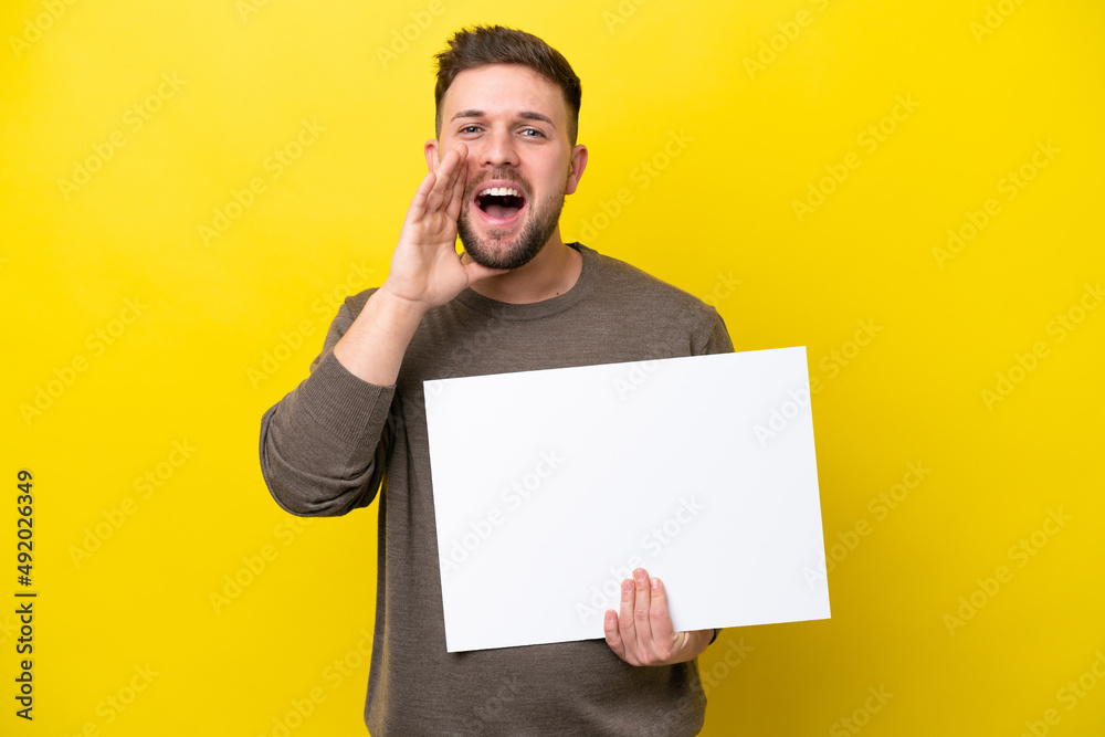 Young caucasian man isolated on yellow background holding an empty placard and shouting Young caucasian man isolated on yellow background holding an empty placard and shouting