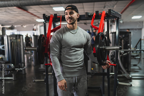 Chico joven musculoso con chándal gris y gorra negra posando en un gimnasio rodeado de maquinas de musculación 