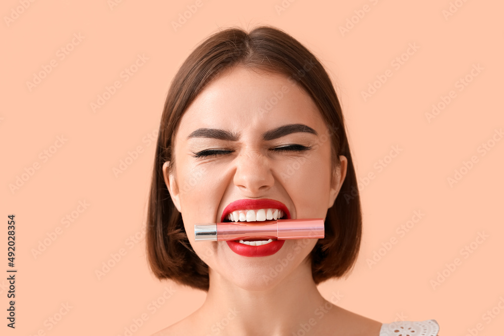 Emotional young woman with red lips holding lipstick in mouth on color background