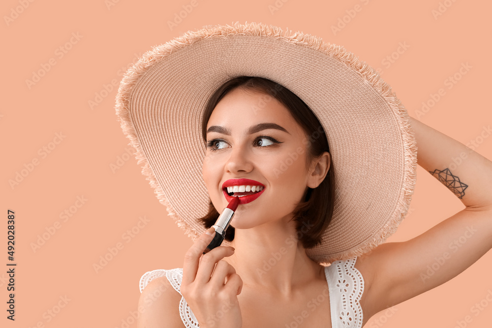 Happy woman in hat adjusting red lipstick on beige background - obrazy, fototapety, plakaty 