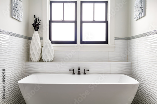 Contemporary Freestanding Bathtub. White bathroom with white custom tile and black faucet. Modern decor with windows.