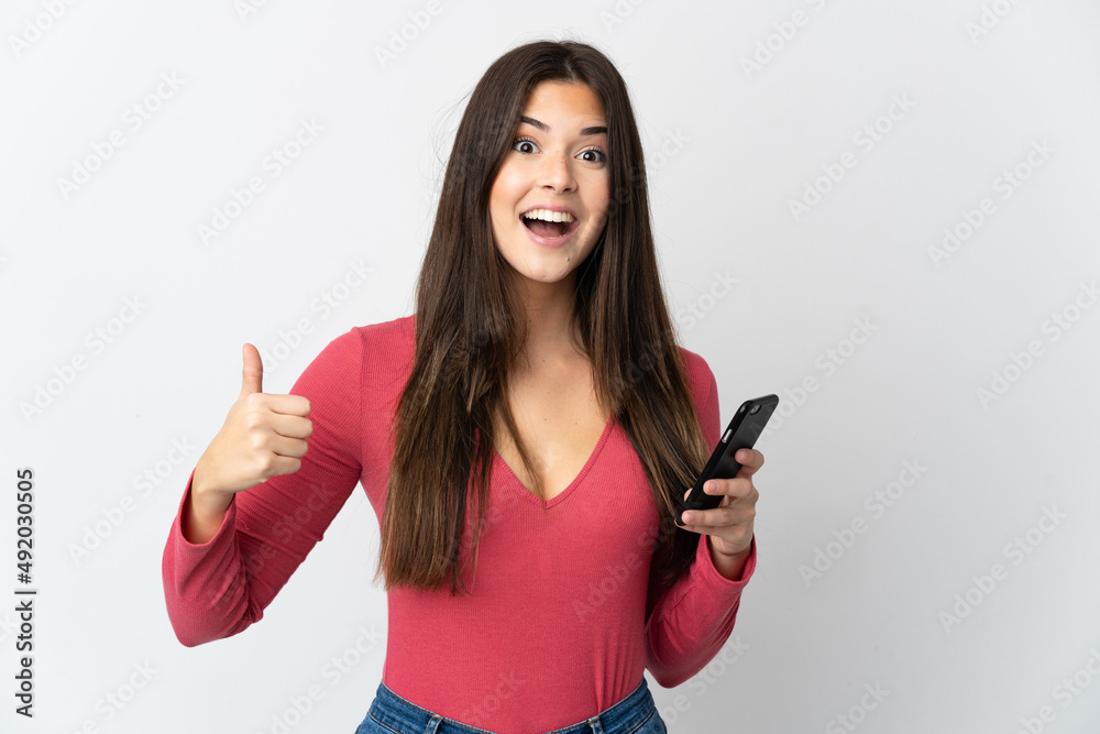 Teenager Brazilian girl isolated on white background using mobile phone while doing thumbs up
