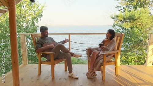 Wallpaper Mural Long shot of Biracial man playing acoustic guitar, his young wife drinking hot tea, sitting on chair on terrace by lake on sunny day in summer, talking Torontodigital.ca