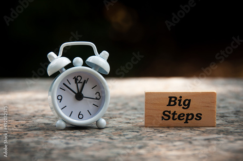 Big steps text on wooden block with white alarm clock and blurred park background. Conceptual
