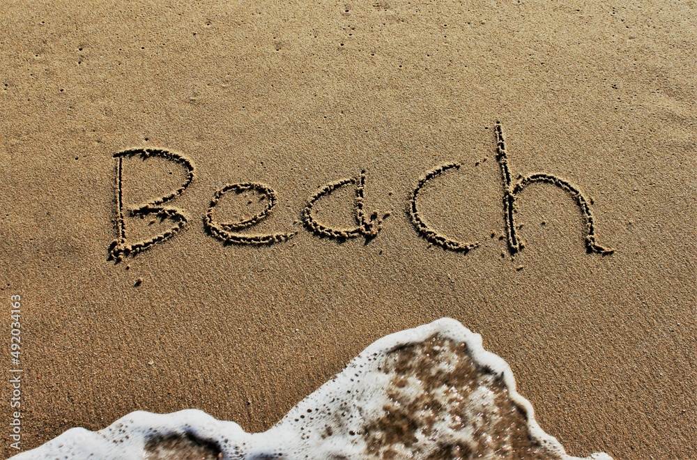 Fototapeta premium On the beach, the words Beach are carved in letters in the smooth sand, which is washed away by a wave