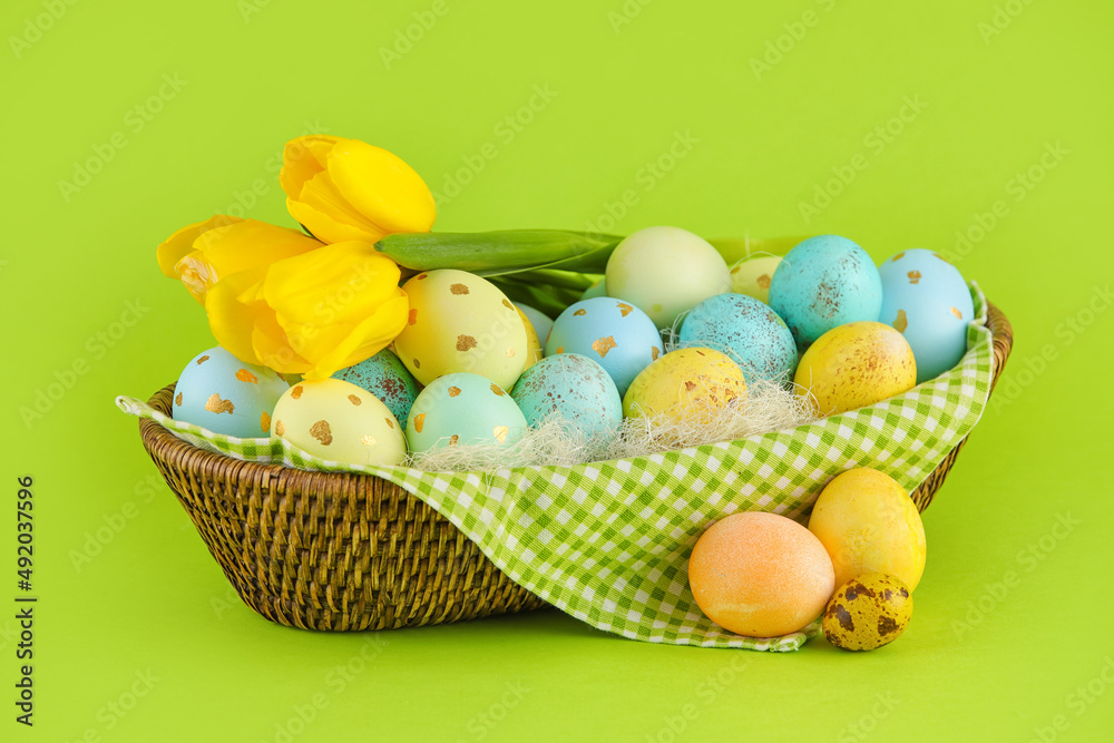 Wicker basket of painted Easter eggs and flowers on green background