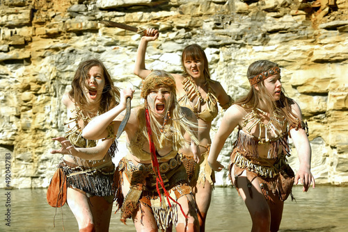 A group of woman dress up as a Neanderthal warriors. Their bodies and faces are covered with mud and dirt. Their facial expression is filled with anger as they wade through water. 