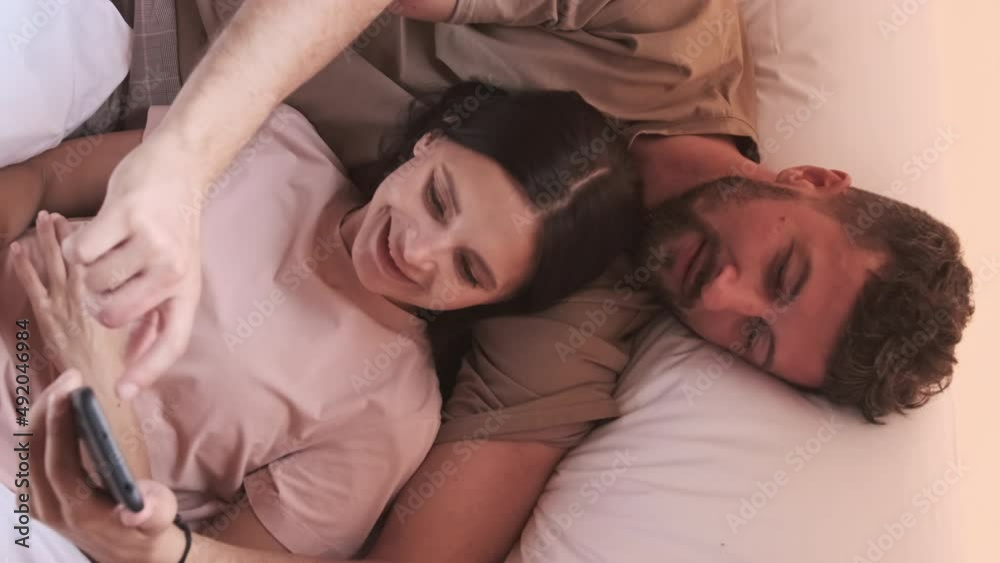 Top view of young dark-haired Biracial woman and bearded husband lying in bed in hotel room, using smartphone and talking in morning Top view of young dark-haired Biracial woman and bearded husband lying in bed in hotel room, using smartphone and talking in morning
