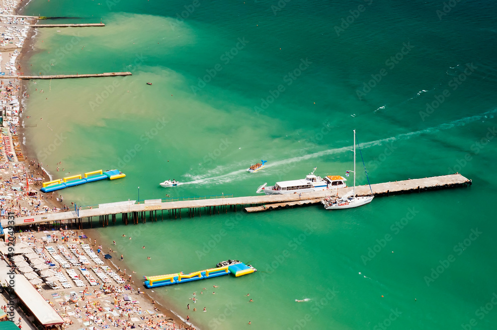 View from a height of the city beach Sudak in the Crimea. Summer vacation at sea. Pier with boats and turquoise water