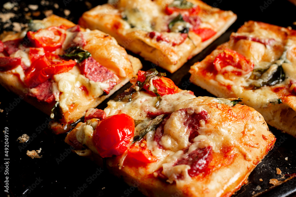 Sliced square pieces of pizza, pie, with tomatoes, sausage, cheese cooked in the oven on a black baking sheet. Sliced square pieces of pizza, pie, with tomatoes, sausage, cheese cooked in the oven on a black baking sheet.