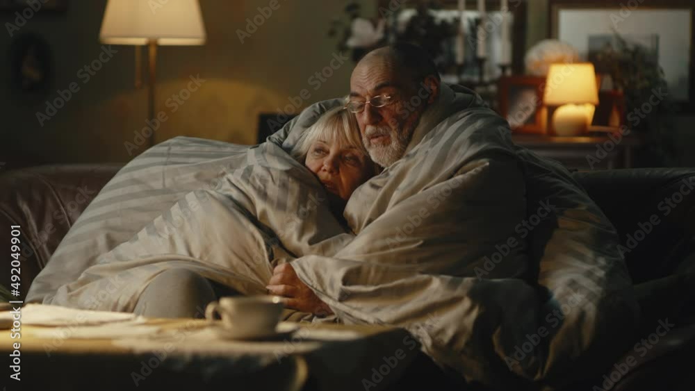 Upset senior husband and wife embracing each other and speaking under warm duvet while sitting on sofa in cold apartment during gas crisis Upset senior husband and wife embracing each other and speaking under warm duvet while sitting on sofa in cold apartment during gas crisis