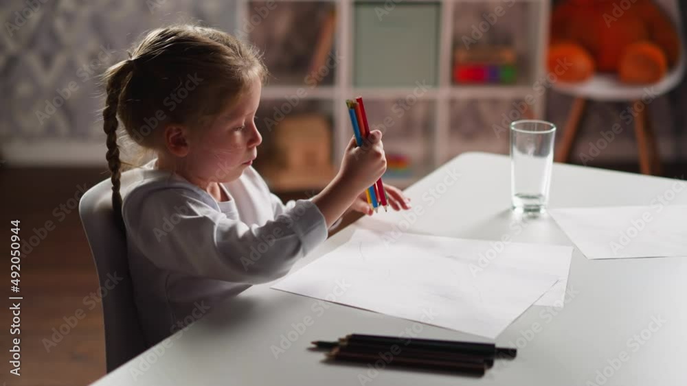 Inspired little artist in white shirt gathers different crayons in bunch to draw on paper sheet sitting at table in light children room slow motion Inspired little artist in white shirt gathers different crayons in bunch to draw on paper sheet sitting at table in light children room slow motion