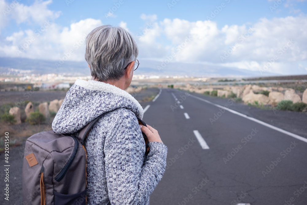 Fototapeta premium Back view of alone senior woman walking outdoor wearing backpack looking at mountain landscape. Elderly mature lady in empty road, mountain and blue sky on background