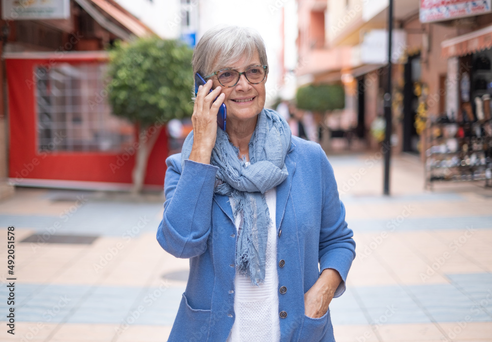 Attractive relaxed mature adult Caucasian woman with gray hair walking in city street holding cellphone in hand. Beautiful senior woman blue dressed enjoys free time talking with smart phone