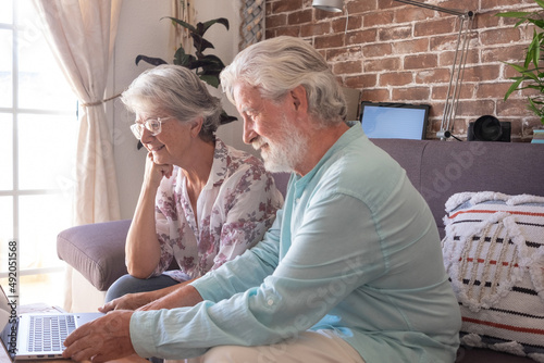 Two caucasian mature modern senior people sitting on sofa at home using laptop computer. Elderly couple gray haired relaxing and smiling together looking at same laptop. Brick wall background