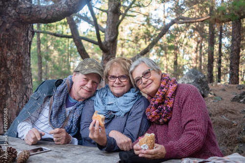 Happy group of seniors people looking at camera holding a sandwich in the hands. Sitting outdoor at a wooden table in the mountain picnic area spending time together enjoying freedom and retirement
