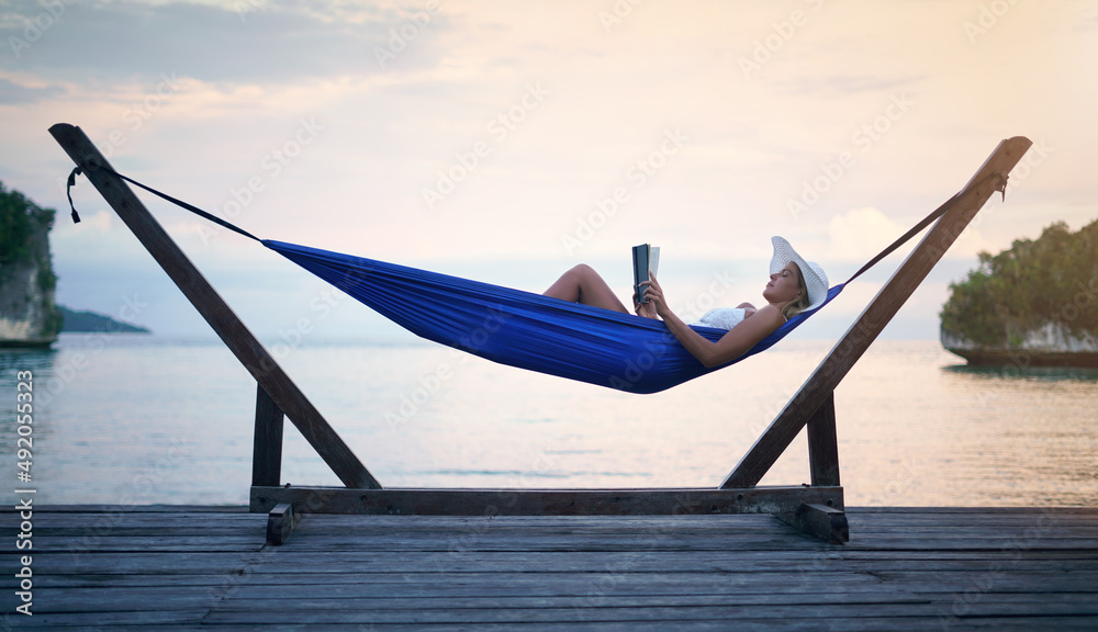 Fototapeta premium Shh, Im on vacation mode. Full length shot of an attractive young woman sitting alone in a hammock and reading a book during a vacation.
