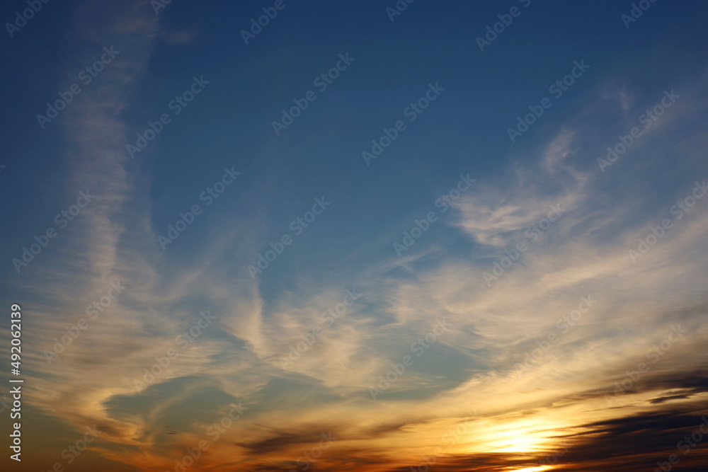 Sunset on a blue sky, sun is shining through the dark and orange clouds. Picturesque landscape for background with soft colors