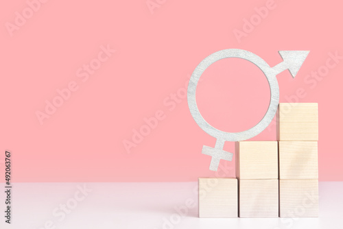 Fototapeta Naklejka Na Ścianę i Meble -  The concept of gender equality, mockup on a pink background with space for text. The symbol of gender equality in silver color stands on wooden cubes arranged in the form of a pyramid ladder
