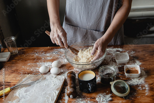a woman in a grey apron prepares dough with wooden shovel in a transparent bowl on a wooden table. On the table are eggs, spices and a glass of milk. 