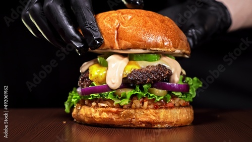 Craft burger is cooking on black background. Consist: red sauce salsa, lettuce, red onion, pickle, cheese, chilli green pepper, air bun and marbled meat beef. Not made ideal. Loving hand made.