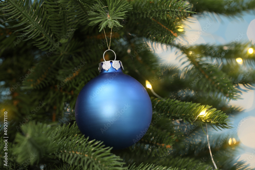 Fototapeta premium Beautiful blue bauble hanging on Christmas tree against blurred festive lights, closeup