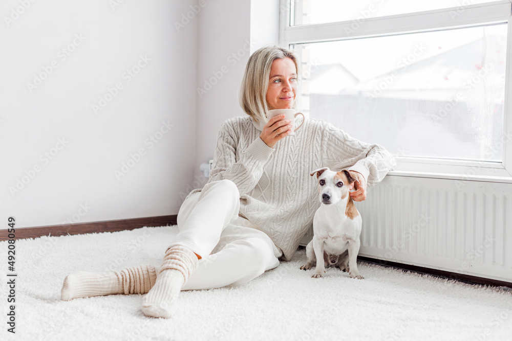 Happy woman in a cozy sweater with dog at home next to the radiator