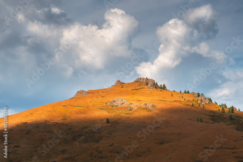 Amazing mountain landscape with cone rock in golden sunlight under beautiful white clouds. Nature background of rocky mountain and green cedar forest. Colorful sunny backdrop with high rocky mountain