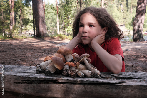 Portrait of cute curly boy sitting at wooden table with a heap of mushrooms on it on sunny summer day.