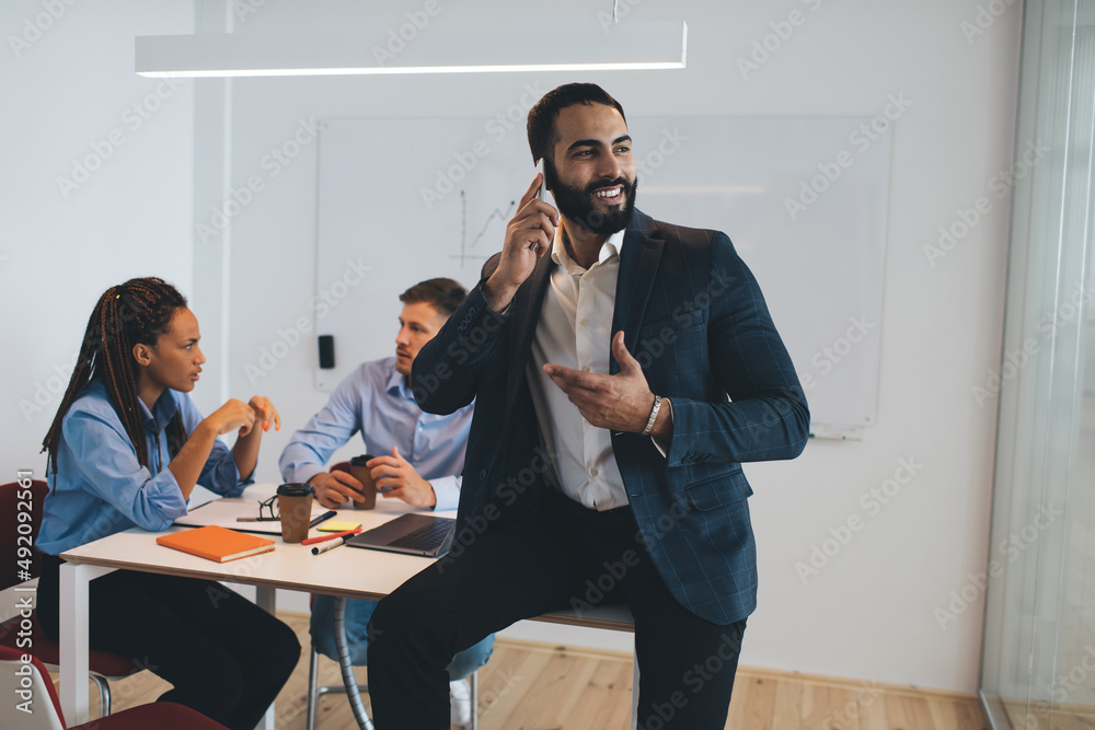 Successful businessman in formal clothes smiling while making cellphone conversation during work time, cheerful Middle Eastern male boss enjoying smartphone communication in office interior Successful businessman in formal clothes smiling while making cellphone conversation during work time, cheerful Middle Eastern male boss enjoying smartphone communication in office interior