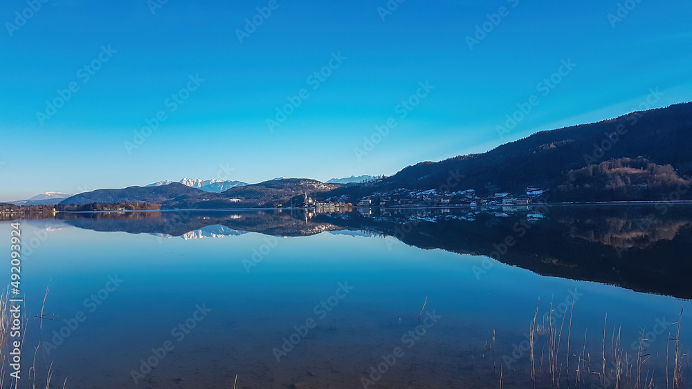 View on Maria Worth and Woerthersee from Poertschach in Carinthia, Austria. The calm surface of the lake is reflecting the mountains, sunbeams and clouds. Village with church located at the lake side View on Maria Worth and Woerthersee from Poertschach in Carinthia, Austria. The calm surface of the lake is reflecting the mountains, sunbeams and clouds. Village with church located at the lake side