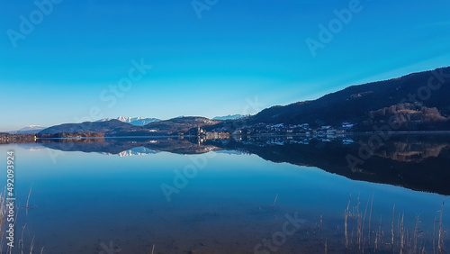 View on Maria Worth and Woerthersee from Poertschach in Carinthia, Austria. The calm surface of the lake is reflecting the mountains, sunbeams and clouds. Village with church located at the lake side