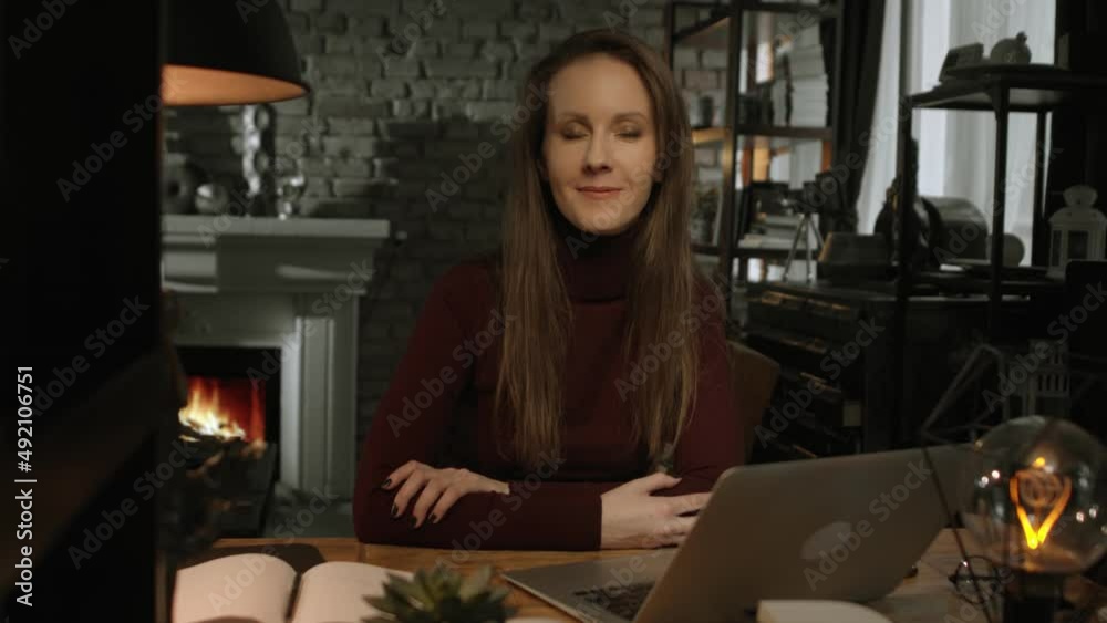 Home office portrait - happy white woman working from home with laptop computer. Sitting at desk in dark living room in front of fireplace in Winter. Home office portrait - happy white woman working from home with laptop computer. Sitting at desk in dark living room in front of fireplace in Winter.