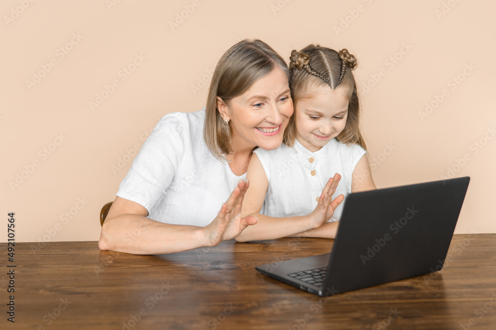 The girl and her grandmother talk with video communication on a laptop and wave their hands like a greeting. Isolated portrait on a beige background. The girl and her grandmother talk with video communication on a laptop and wave their hands like a greeting. Isolated portrait on a beige background.