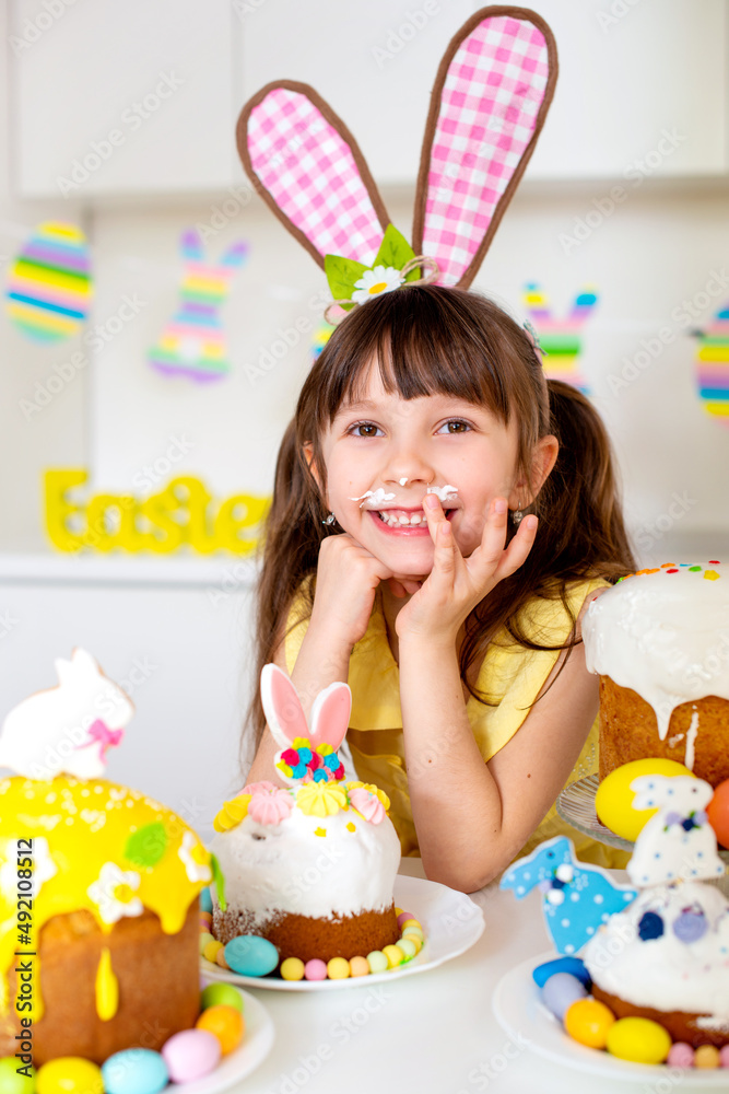 A cute little girl with bunny ears prepares an Easter cake and painted eggs. Religious holiday A cute little girl with bunny ears prepares an Easter cake and painted eggs. Religious holiday