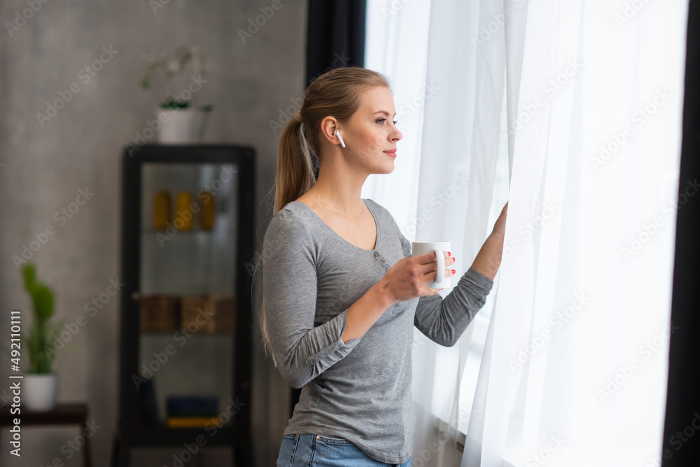 Young teenage girl is standing in front of the window and looking into it. Blond woman at home. Young teenage girl is standing in front of the window and looking into it. Blond woman at home.