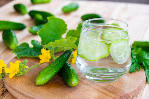 Glass with cucumber water on wooden board with fresh cucumbers around. Homemade lemonade of fresh cucumbers for summer refreshment, diet and detox.