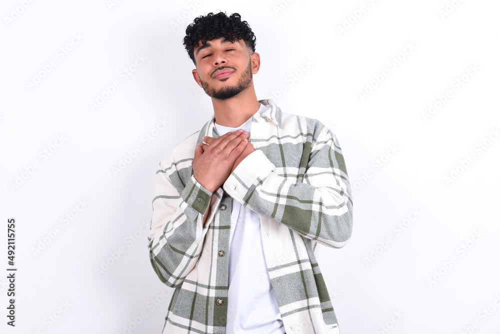 young arab man with curly hair wearing overshirt closes eyes and keeps hands on chest near heart, expresses sincere emotions, being kind hearted and honest. Body language and real feelings concept. young arab man with curly hair wearing overshirt closes eyes and keeps hands on chest near heart, expresses sincere emotions, being kind hearted and honest. Body language and real feelings concept.