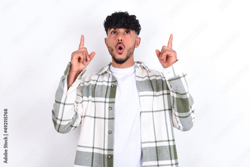 young arab man with curly hair wearing overshirt over white background being amazed and surprised looking and pointing up with fingers showing something strange. young arab man with curly hair wearing overshirt over white background being amazed and surprised looking and pointing up with fingers showing something strange.