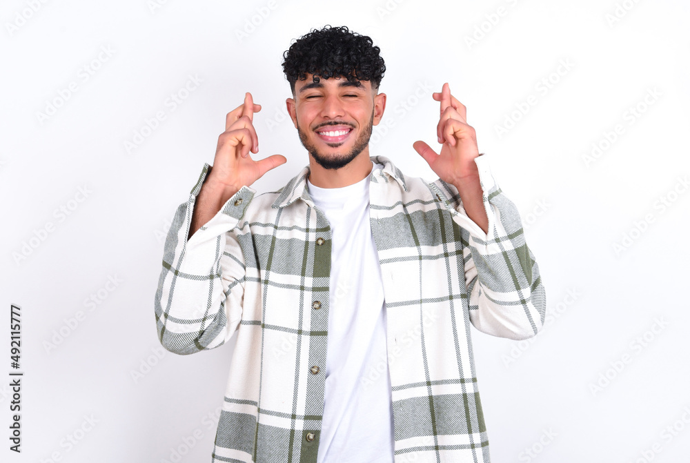 Joyful young arab man with curly hair wearing overshirt over white background clenches teeth, raises fingers crossed, makes desirable wish, waits for good news, I have to win. Joyful young arab man with curly hair wearing overshirt over white background clenches teeth, raises fingers crossed, makes desirable wish, waits for good news, I have to win.