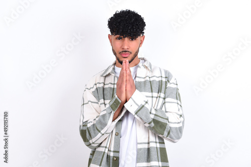 young arab man with curly hair wearing overshirt over white background keeps palms pressed together in front of her having regretful look, asking for forgiveness. Forgive me please.