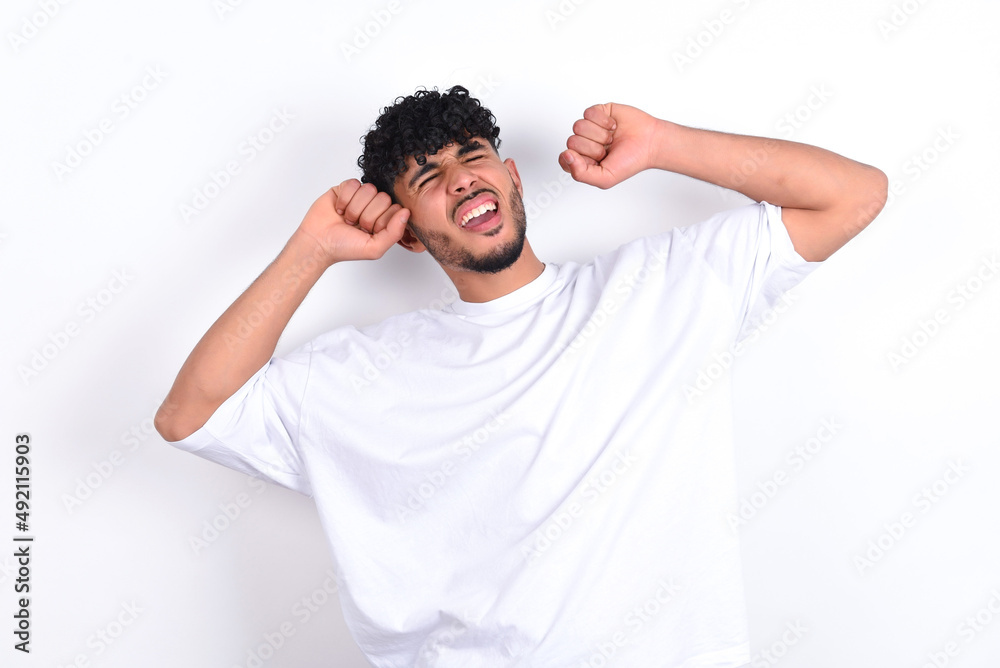 young arab man with curly hair wearing white t-shirt over white background relaxing and stretching, arms and hands behind head and neck smiling happy young arab man with curly hair wearing white t-shirt over white background relaxing and stretching, arms and hands behind head and neck smiling happy